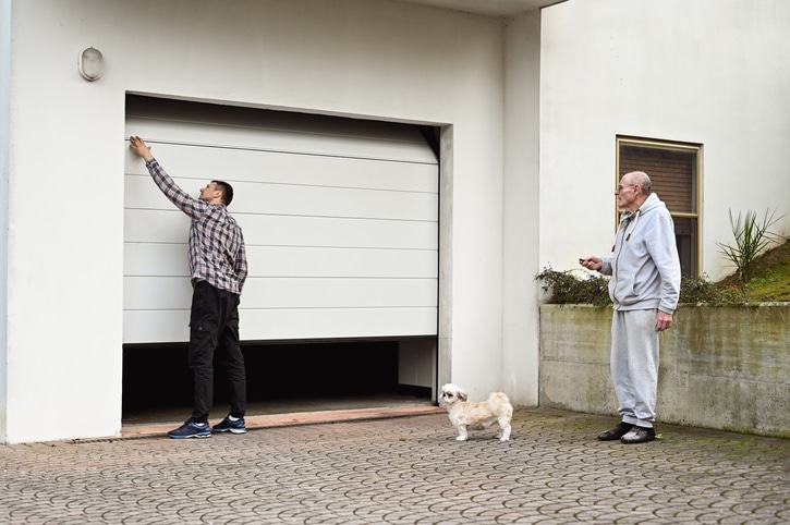 A man opens a garage door while another man stands nearby holding a remote, with a small dog standing on the driveway.