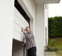 A man in a plaid shirt is manually opening a white garage door attached to a modern house.
