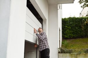 A man in a plaid shirt is manually opening a white garage door attached to a modern house.