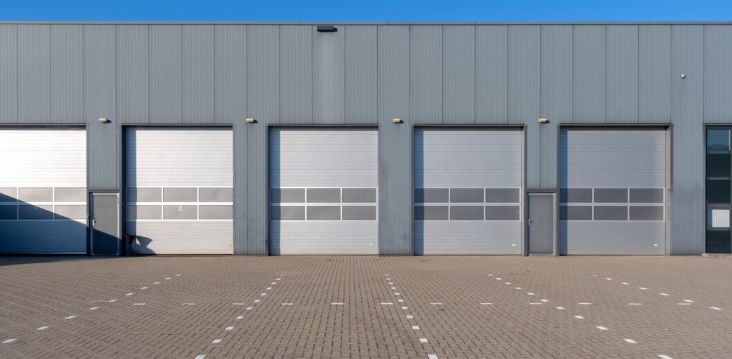 A row of closed industrial garage doors on a gray warehouse building, with an empty paved lot in front and clear blue sky above.