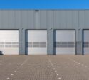 A row of closed industrial garage doors on a gray warehouse building, with an empty paved lot in front and clear blue sky above.