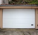 A closed white garage door set between stone walls with a tiled roof above and a concrete driveway in front.