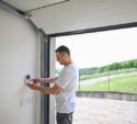A man stands inside a garage, pressing buttons on a wall-mounted keypad, with the garage door open to an outdoor view.