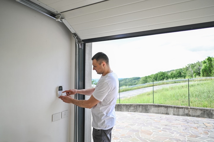 A man stands inside a garage, pressing buttons on a wall-mounted keypad, with the garage door open to an outdoor view.