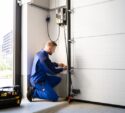 Technician in blue overalls kneels to repair or install the control panel of an automatic garage door, with a tool bag placed nearby.