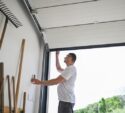 A person in a white t-shirt opens a garage door while standing next to a wall with gardening tools.
