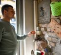 A man in a green sweatshirt stands indoors by a brick wall with tools and a thermostat, reaching toward the thermostat.