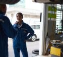 Two auto mechanics in blue uniforms are talking in a garage, with equipment and a car visible around them.