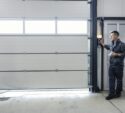 A man in work clothes stands by a control panel, operating a large white industrial garage door in a workshop or warehouse setting.