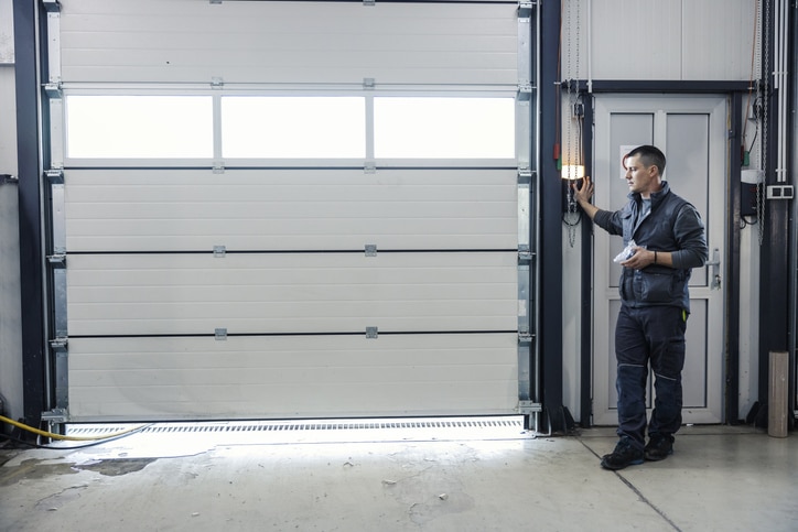 A man in work clothes stands by a control panel, operating a large white industrial garage door in a workshop or warehouse setting.