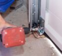 A person uses a power drill to install or repair a metal bracket at the base of a garage door track on a concrete floor.