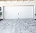 Closed white garage door with two handles, bordered by two black lantern lights, shrubs, and flowering plants on either side, with patterned stone driveway in front.
