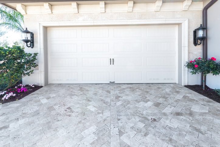 Closed white garage door with two handles, bordered by two black lantern lights, shrubs, and flowering plants on either side, with patterned stone driveway in front.