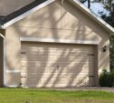 A beige single-car garage with a closed sectional door attached to a house, surrounded by greenery and a paved driveway.