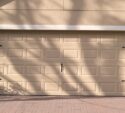 Closed beige garage door with rectangular panels, black decorative hardware, and shadows of tree branches on the surface. Brick-patterned driveway in front.