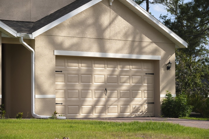 A beige single-car garage with a closed sectional door attached to a house, surrounded by greenery and a paved driveway.