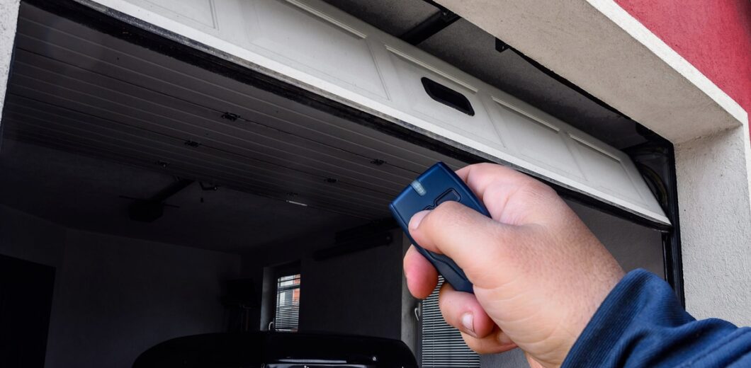 A person uses a remote control to open a white garage door, revealing a parked car inside the garage.