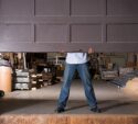 A person in jeans and a shirt lifts a large garage door in a warehouse filled with various stacked materials and equipment.