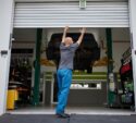 A person in blue pants opens a garage door, with a car elevated on a lift inside an automotive workshop.