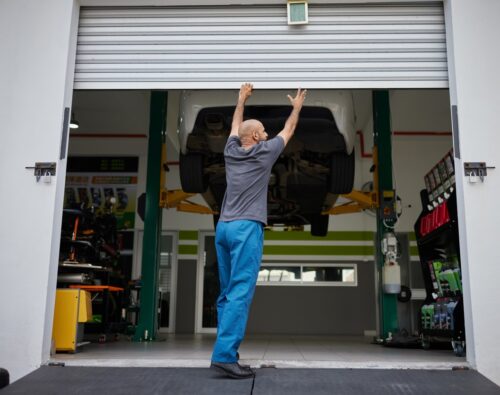 A person in blue pants opens a garage door, with a car elevated on a lift inside an automotive workshop.