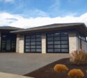 Modern house with two large glass garage doors, stone accents, and a landscaped front yard with bushes and a concrete driveway under a partly cloudy sky.