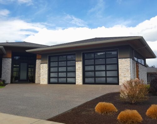 Modern house with two large glass garage doors, stone accents, and a landscaped front yard with bushes and a concrete driveway under a partly cloudy sky.