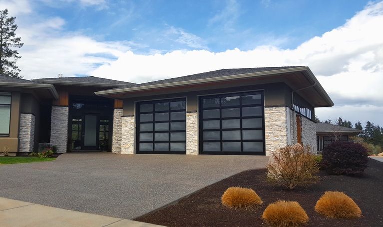 Modern house with two large glass garage doors, stone accents, and a landscaped front yard with bushes and a concrete driveway under a partly cloudy sky.