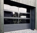 A modern garage door with horizontal panels and large dark glass windows set in a wooden building, with cobblestone pavement in front.