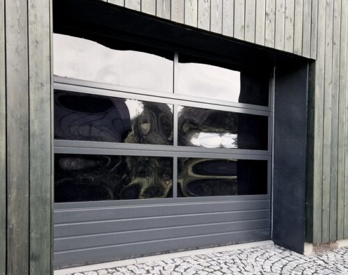 A modern garage door with horizontal panels and large dark glass windows set in a wooden building, with cobblestone pavement in front.