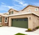 Two-story beige suburban house with a green garage door, stone accents, manicured lawn, and clear blue sky in the background.