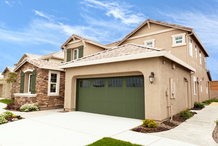 Two-story beige suburban house with a green garage door, stone accents, manicured lawn, and clear blue sky in the background.