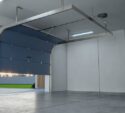 Interior of an empty garage with a partially open blue overhead door, gray concrete floor, and a door on the right wall.