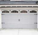 Closed gray garage door with decorative handles, window panels at the top, and stone accents on the sides of a residential house exterior.