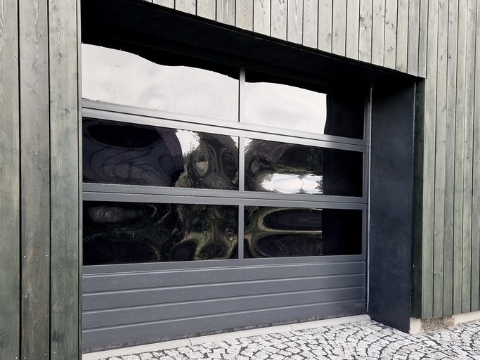A modern garage door with horizontal panels and large dark glass windows set in a wooden building, with cobblestone pavement in front.
