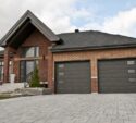 A modern brick house with two dark garage doors, a paved driveway, front steps, and minimal landscaping under a partly cloudy sky.