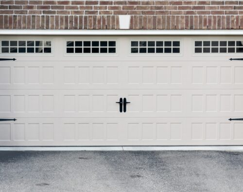 Closed beige garage door with rectangular panels and three small windows at the top, set in a brick and stone exterior wall.