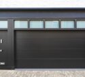 Modern black garage door with four rectangular windows above and an adjacent black entrance door, set in a white brick building with a stone driveway.