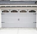 A closed, double-car garage door with vertical panel design, decorative windows, and black hardware on a modern house with stone accents.
