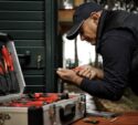 A person in a cap and vest uses a screwdriver to work on a lock, with an open toolbox of various tools nearby.