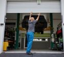 A person in blue pants opens a garage door in an auto repair shop with a car lifted on a hydraulic platform inside.