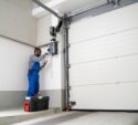 A person wearing blue coveralls repairs or installs a garage door opener using tools, with a toolbox placed on the floor nearby.