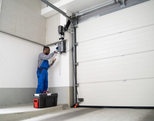 A person wearing blue coveralls repairs or installs a garage door opener using tools, with a toolbox placed on the floor nearby.