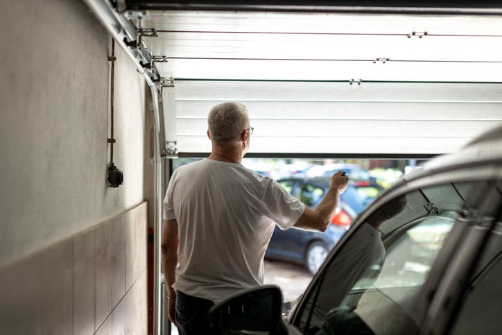 A man in a white t-shirt stands inside a garage, holding a remote and opening the garage door, with cars visible outside.