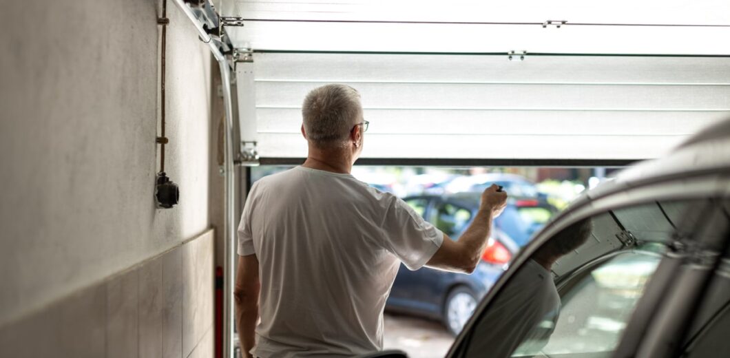 A man with short gray hair stands inside a garage, holding a remote control while opening the garage door. A car is parked inside, and other vehicles are visible outside.