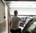 A man with short gray hair stands inside a garage, holding a remote control while opening the garage door. A car is parked inside, and other vehicles are visible outside.
