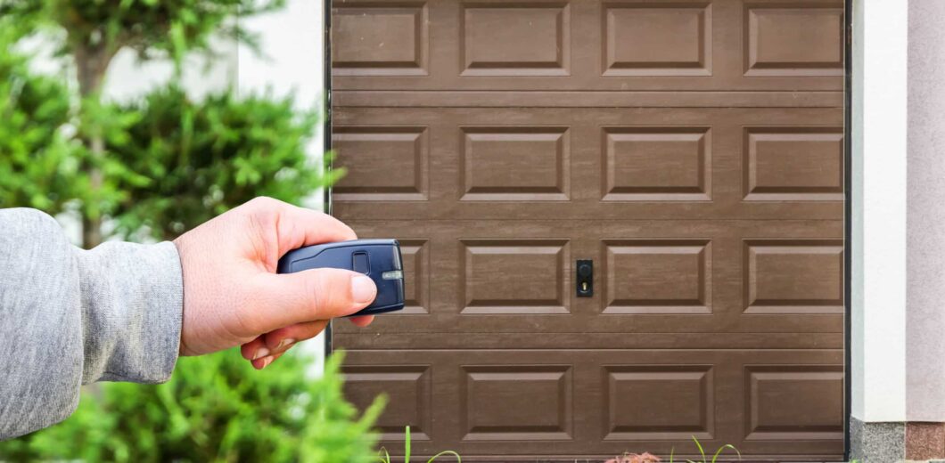 A person’s hand holds a remote control in front of a closed brown garage door, with greenery visible in the foreground.