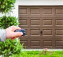 A person’s hand holds a remote control in front of a closed brown garage door, with greenery visible in the foreground.