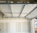 Partially open white garage door inside an unfinished garage with exposed drywall. Exterior view shows houses and construction materials.