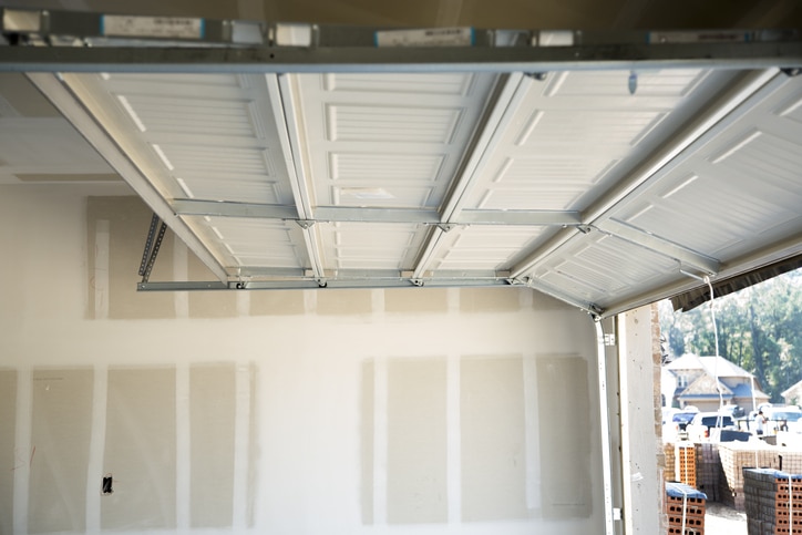 Partially open white garage door inside an unfinished garage with exposed drywall. Exterior view shows houses and construction materials.