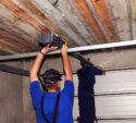 Two workers in blue uniforms install or repair a garage door opener mechanism on the ceiling of a garage with exposed wooden beams.